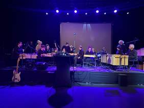 Pokaż zdjęcie: Na zdjęciu widać orkiestrę perkusyjną Gamelan z PSONI Szczecin podczas występu scenicznego. Grupa składa się z kilkunastu muzyków w różnym wieku, siedzących przy tradycyjnych instrumentach perkusyjnych ustawionych w półokręgu na podświetlonej scenie. W centralnym punkcie znajduje się wolnostojący mikrofon, a przed nim — stół z konsoletą dźwiękową. W tle widać ciemne kurtyny i duży ekran bez wyświetlanej treści. Scena oświetlona jest niebiesko-fioletowym światłem, które tworzy nastrojowy klimat. Po lewej stronie oparta jest elektryczna gitara, a po prawej – widoczna jest perkusja marki Yamaha. Artyści są skupieni i przygotowani do gry, co tworzy atmosferę oczekiwania na rozpoczęcie koncertu.