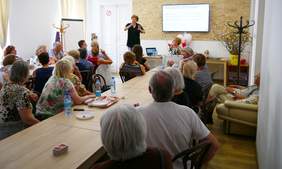 Pokaż zdjęcie: Grupa osób siedząca przy stolikach patrząca na tłumacza języka migowego