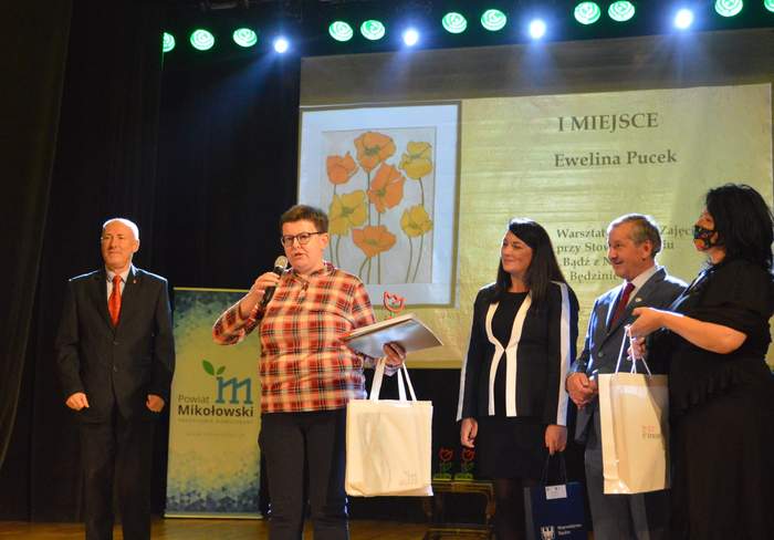 Pokaż zdjęcie: na scenie stoi kobieta i mówi do mikrofonu w drugiej ręce trzyma nagrody i dyplomy obok niej po prawej stronie stoją 3 osoby a po lewej mężczyzna