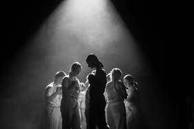 Pokaż zdjęcie: zdjęcie w kolorze czarno białym, przedstawia osoby zebrane w kręgu, trzymają w rękach przedmiot nieokreślony ale puszysty, zwrok ich jest skierowany ku dołowi 