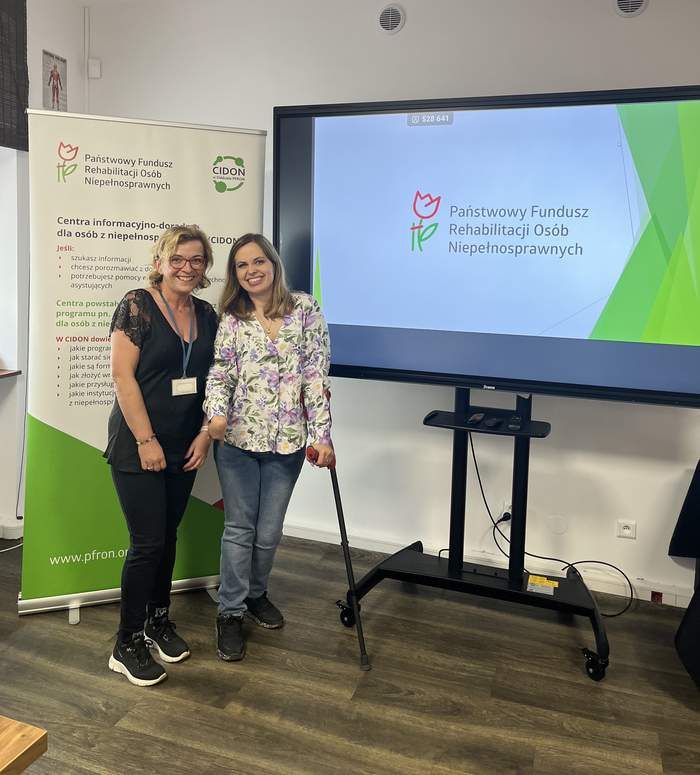 Pokaż zdjęcie: Dwie kobiety stoją obok siebie na tle rollupu PFRON i ekranu z logo PFRON i są uśmiechnięte oraz zadowolone