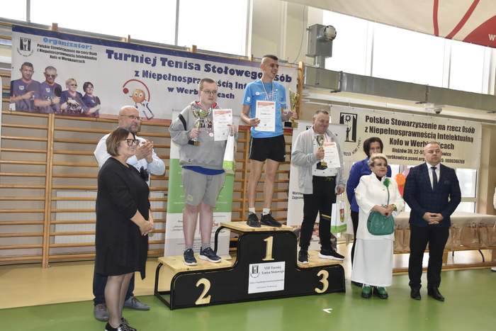 Pokaż zdjęcie: sala gimnastyczna, zdjęcie osób stojących na podium, w rękach trzymają dyplomy, na szyi mają zawieszone medale. Obok podium stoją trzy kobiety i jeden mężczyzna.