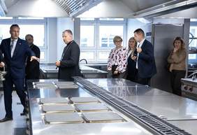 Pokaż zdjęcie: Grupa przedstawicieli władz i duchowieństwa ogląda nowoczesną, stalową kuchnię przemysłową, zapoznając się z jej wyposażeniem oraz warunkami pracy podczas wizyty w Zakładzie Aktywności Zawodowej w Sokółce.