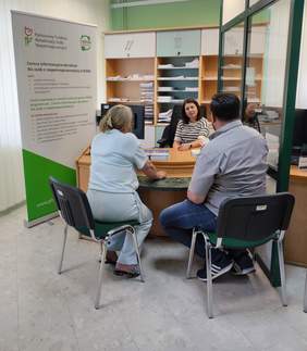 Pokaż zdjęcie: Kobieta z PFRON w paskowanej bluzce prowadzi indywidualną konsultację z dwiema osobami – kobietą w jasnym stroju i mężczyzną w szarej koszuli. Siedzą przy biurku w urzędzie, obok znajduje się roll-up z informacjami o Centrach Informacyjno-Doradczych dla Osób z Niepełnosprawnościami. W tle półki z dokumentami.