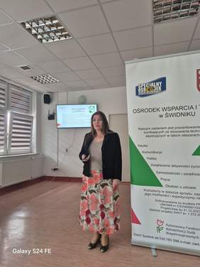 Pokaż zdjęcie: Trwa prezentacja multimedialna, prelegentka stoi przy rollupie, w tle ekran Z prezentacją.