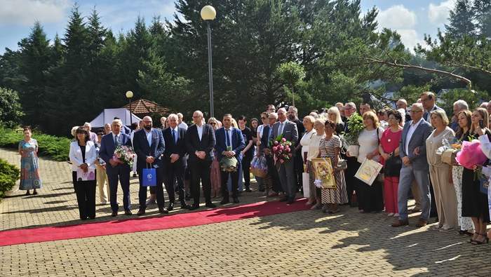 Pokaż zdjęcie: Duża grupa ludzi w eleganckich strojach stoi na zewnątrz na brukowanym placu, niektórzy trzymają kwiaty i obrazy. Czerwony dywan prowadzi przez środek, w tle drzewa.