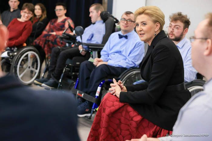 Pokaż zdjęcie: Pośród 10 osób siedzących na wózkach siedzi Pierwsza Dama Agata Kornhauer-Duda, ubrana w długą czerwoną suknię i czarną marynarkę z paskiem.