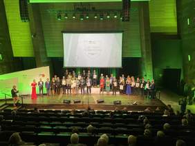 Pokaż zdjęcie: Na scenie w oddali stoi wielu uśmiechniętych mężczyzn i kobiet ubranych na galowo. Trzymają w rękach dyplomy puchary, na ich szyjach wiszą medale. Część z nich stoi na specjalnie przygotowanych podwyższeniach świadczących o zajętych miejscac