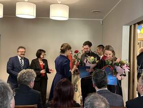 Pokaż zdjęcie: Kobieta w eleganckiej sukience wręcza kwiaty i dyplomy pracownikom. Obok stoi kobieta w eleganckiej garsonce i mężczyzna w garniturze. Przed nimi siedzą goście.