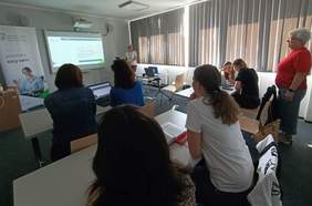 Pokaż zdjęcie: Grupa ośmiu osób znajduje się w sali szkoleniowej. Po prawej stronie sali stoi kobieta prowadząca szkolenie. Na dużym ekranie wyświetlana jest prezentacja. Po lewej stronie ustawiony jest baner z logo PFRON.