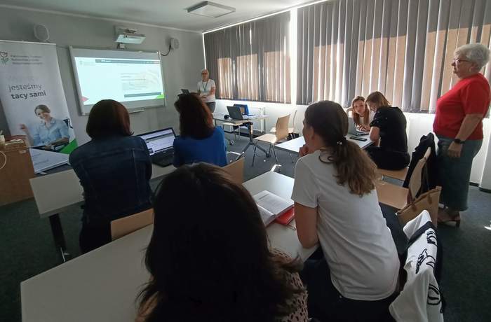 Pokaż zdjęcie: Grupa ośmiu osób znajduje się w sali szkoleniowej. Po prawej stronie sali stoi kobieta prowadząca szkolenie. Na dużym ekranie wyświetlana jest prezentacja. Po lewej stronie ustawiony jest baner z logo PFRON.