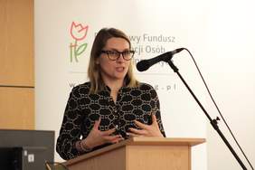 Pokaż zdjęcie: Przy mównicy kobieta ubrana w sukienkę w drobny print. Gestykuluje rękami