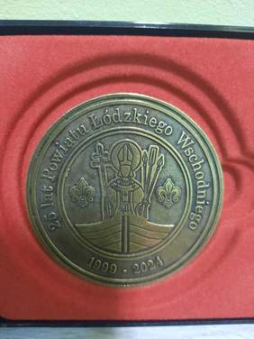 Pokaż zdjęcie: Na czerwonym tle leży medal z wizerunkiem Świętego Wojciecha. Na otoczce odznaczenia napisane jest: 25 lat Powiatu Łódzkiego Wschodniego 1999-2024. 