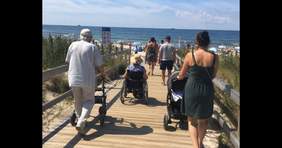 Pokaż zdjęcie: Na plażę prowadzi utwardzona kładka. W stronę morza idzie grupa osób. Są wśród nich osoby starsze, pani na wózku inwalidzkim i kobieta prowadząca wózek z dzieckiem. Jest piękna pogoda. 