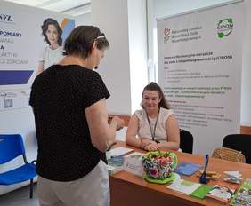 Pokaż zdjęcie: Przy biurku na którym leżą ulotki rozmawiają dwie kobiety. 