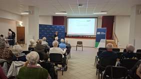 Kobieta stoi z mikrofonem w ręku, grupa starszych osób siedzi, w tle prezentacja na dużym ekranie