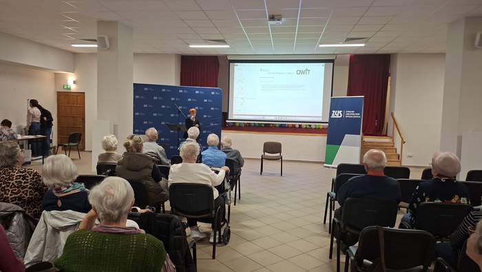 Kobieta stoi z mikrofonem w ręku, grupa starszych osób siedzi, w tle prezentacja na dużym ekranie