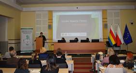 Pokaż zdjęcie: Zdjęcie przedstawia salę konferencyjną z uczestnikami siedzącymi przy stołach. Przy mównicy stoi kobieta w okularach wygłaszająca prelekcję, a za nią na ekranie wyświetlona jest prezentacja zatytułowana Ośrodek Wsparcia i Testów w Białymstoku. Po prawe stronie stoją trzy flagi: Polski, województwa podlaskiego i Unii Europejskiej. Po lewej stronie widoczny jest baner Regionalnego Ośrodka Polityki Społecznej w Białymstoku. Przy stole prezydialnym siedzą trzy osoby, korzystające z laptopów.