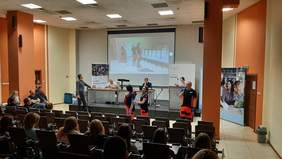 Zdjecie sali z uczestnikami, siedzącymi tyłem, na  scianie wisi ekran, a z przodu stoją ratownicy medyczni