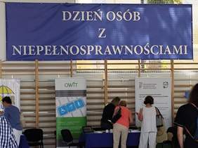 Pokaż zdjęcie: Duża sala, w której odbywa się wydarzenie. Na ścianie transparent z napisem Dzień Osób z Niepełnosprawnością. Pod transparentem stoiska informacyjne do których podchodzą uczestnicy.