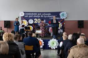 Pokaż zdjęcie: Zdjęcie z tyłu sali w kierunku sceny, na której stoją trzy kobiety oraz mężczyzna w garniturze przy mównicy.