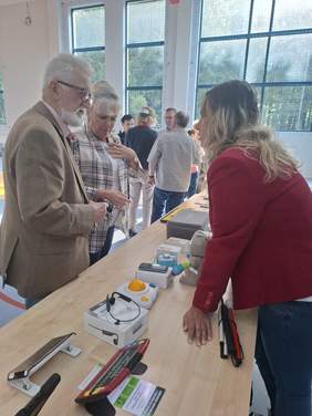 Pokaż zdjęcie: Starszy mężczyzna i kobieta rozmawiają z młodszą kobietą stojącą za stołem. Na stole rozłożone są różne urządzenia – technologie wspomagające dla osób z niepełnosprawnościami.