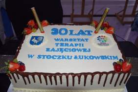 Pokaż zdjęcie: Biały kwadratowy tort z racami i napisem 30 lat Warsztatu Terapii Zajęciowej w Stoczku Łukowskim.