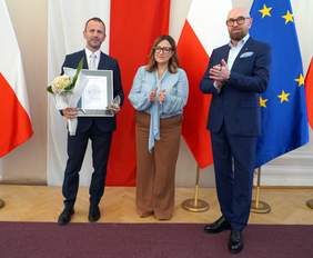 Pokaż zdjęcie: Na tle flagi Polski oraz Unii Europejskiej pozują trzy osoby, jedna z osób ma dyplom i kwiaty w ręku.