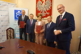 Pokaż zdjęcie: Przy stole stoi pięć osób, w rękach trzymaja teczki z dokumentami