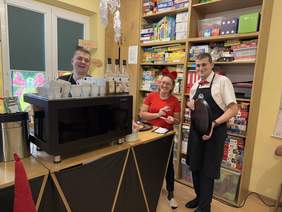 Pokaż zdjęcie: Trzy osoby w stoją przy dużym ekspresie do kawy w sali pełnej gier planszowych. Mężczyzna z lewej uśmiecha się, kobieta w czerwieni trzyma notatki, a kelner w muszce trzyma tacę. W tle półki z grami, kolorowe dekoracje na oknach.