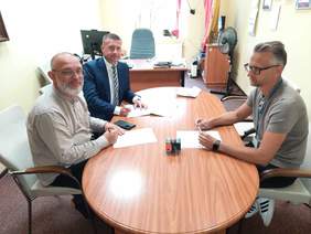 Pokaż zdjęcie: Przy owalnym stole siedzi trzech panów. Dwóch z nich uśmiecha się i patrzy w obiektyw aparatu. Na stole leżą dokumenty, pieczątki, telefon. 