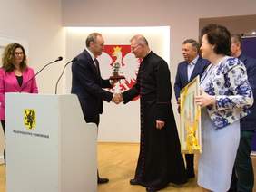 Pokaż zdjęcie: Mężczyzna w garniturze w trzyma nagrodę i ściska dłoń księdza ubranego w sutannę. Obok stoją kobiety jedna trzyma rysunek w ramie. Przed nimi mównica z napisem Województwo Pomorskie. W tle godło polskie.
