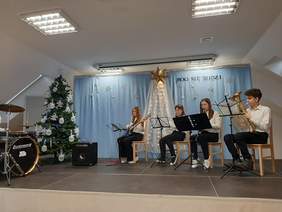 Pokaż zdjęcie: Na scenie siedzą dzieci grające na sprzętach: gitarze, flecie, trąbce, perkusji. Na scenie jest świąteczny wystrój, stoi udekorowana na biało choinka, na niebieskiej kotarze przypięto napis Bóg się rodzi , a dookoła napisu są białe gwiazdki.