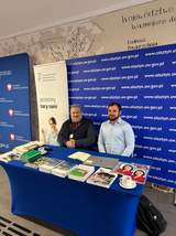 Dwóch mężczyzn przy stoisku informacyjnym PFRON. Na stole znajdują się materiały i publikacje dla uczestników wydarzenia.