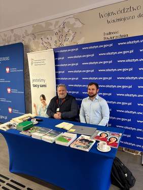 Pokaż zdjęcie: Dwóch mężczyzn przy stoisku informacyjnym PFRON. Na stole znajdują się materiały i publikacje dla uczestników wydarzenia.
