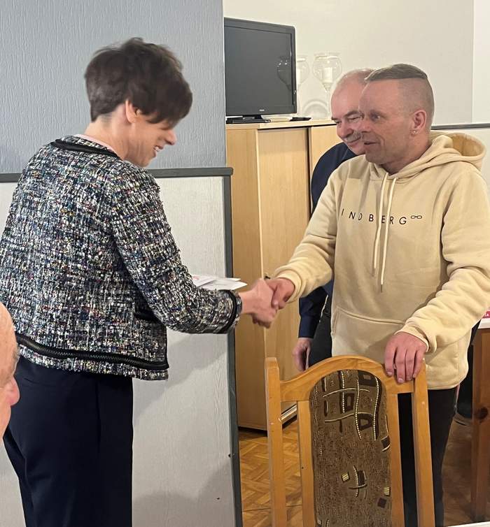 Pokaż zdjęcie: Kobieta w czarno-białym żakiecie gratuluje laureatowi Konkursu Plastycznego „Sztuka Osób Niepełnosprawnych”, podając mu rękę. Laureat, mężczyzna w jasnej bluzie z kapturem, stoi po prawej stronie. W tle widać częściowo zasłoniętą postać innego mężczyzny, jasną ścianę z telewizorem oraz szafkę. Na pierwszym planie po prawej widoczne jest oparcie drewnianego krzesła.