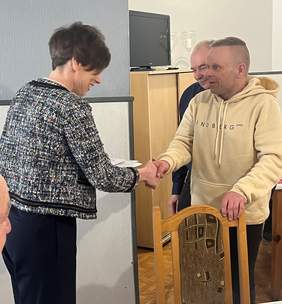 Pokaż zdjęcie: Kobieta w czarno-białym żakiecie gratuluje laureatowi Konkursu Plastycznego „Sztuka Osób Niepełnosprawnych”, podając mu rękę. Laureat, mężczyzna w jasnej bluzie z kapturem, stoi po prawej stronie. W tle widać częściowo zasłoniętą postać innego mężczyzny, jasną ścianę z telewizorem oraz szafkę. Na pierwszym planie po prawej widoczne jest oparcie drewnianego krzesła.