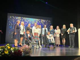 Pokaż zdjęcie: Na scenie Białostockiego Teatru Lalek stoi dziewięć dorosłych osób (nauczycieli) : osiem kobiet i jeden mężczyzna oraz czworo uczniów. Dwie osoby są na wózku inwalidzkim.  Wszyscy biorą udział w występie artystycznym. Mężczyzna stojący przy mównicy,  po prawej stronie sceny, gra na organach i śpiewa. Po lewej stronie sceny leżą dwa bukiety kwiatów, czerwone róże i kolorowe kwiaty.