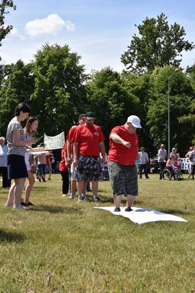 Pokaż zdjęcie: Na trawie leży biały materiał na nim stoi mężczyzna za nim stoi kolejka osób, aby przejść tor przeszkód