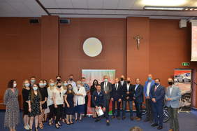 Pokaż zdjęcie: Liczna grupa osób w maseczkach, pozująca do zbiorowej fotografii. W tle baner konkursu i logo uniwersyteckie z łacińską nazwą: Universitas Cardinalis Stephani Wyszyński, Varsoviae. 