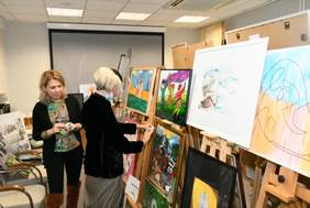 Plan zdjęcia stanowi sala w której na sztalugach ustawione są prace plastyczne. W centrum fotografii stoją członkowie komisji konkursowej.