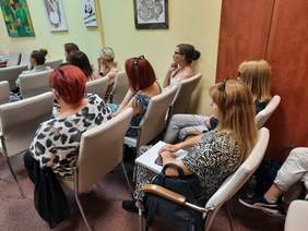 Pokaż zdjęcie: W sali siedzą na krzesłach zasłuchani ludzie. Wszyscy patrzą przed siebie. Na ścianie wisi dużo kolorowych obrazków. 