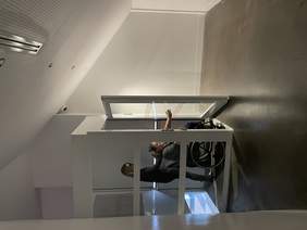 Pokaż zdjęcie: Z dźwigu osobowego wysiadają dwie Kobiety. Jedna porusza się na wózku inwalidzkim druga jest osobą niewidomą w ciemnych okularach. Dźwig i jego drzwi są w kolorze bieli dookoła są białe ściany jest w ciemnym kolorze.