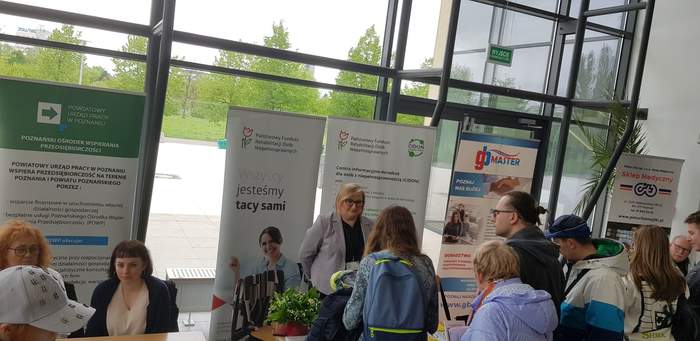 Pokaż zdjęcie: Cztery osoby stoją przy stoisku Oddziału Wielkopolskiego PFRON, przy którym obecna jest kobieta udzielająca informacji. W tle widoczne są banery: PFRON i CIDON. Obok widoczne są stoiska innych wystawców oraz pozostali uczestnicy Targów Pracy.