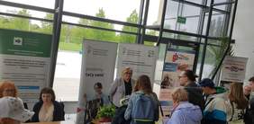 Pokaż zdjęcie: Cztery osoby stoją przy stoisku Oddziału Wielkopolskiego PFRON, przy którym obecna jest kobieta udzielająca informacji. W tle widoczne są banery: PFRON i CIDON. Obok widoczne są stoiska innych wystawców oraz pozostali uczestnicy Targów Pracy.