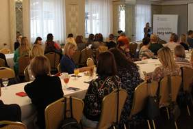 Pokaż zdjęcie: mężczyźni i kobiety patrząna kobietę z mikrofonem w tle prezentacja projektu