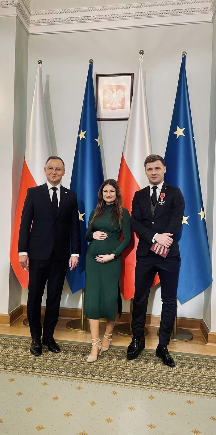 Pokaż zdjęcie: Dwóch mężczyzn i ciężarna kobieta pozują do pamiątkowej fotografii. Jednym z nich jest Prezydent Rzeczypospolitej Polskiej, Andrzej Duda, stojący po lewej stronie. Drugi to odznaczony Krzyżem Oficerskim Orderu Odrodzenia Polski, paraolimpijczyk Piotr Grudzień wraz z małżonką. Za postaciami ustawione naprzemiennie na masztach flagi Polski i Unii Europejskiej.”