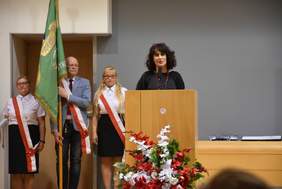 Pokaż zdjęcie: Za mównicą brunetka w czarnej sukience i czerwonymi koralami na szyi. Obok poczet sztandarowy złożony z ubranych na galowo dwóch pań w okularach i pana trzymającego zielony sztandar