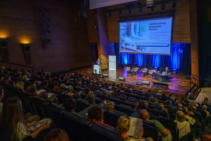 Pokaż zdjęcie: Na auli siedzi dużo ludzi. Na secinie stoi mężczyzna za mównicą obok niego duży ekran na którym wyświetlana jest prezentacja. Na scenie stoi pięć foteli na jednym siedzi mężczyzna.