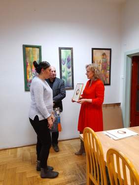 Pokaż zdjęcie: Kobieta, blondynka w czerwonej sukience wręcza dyplom laureatce konkursu, w tle mężczyzna